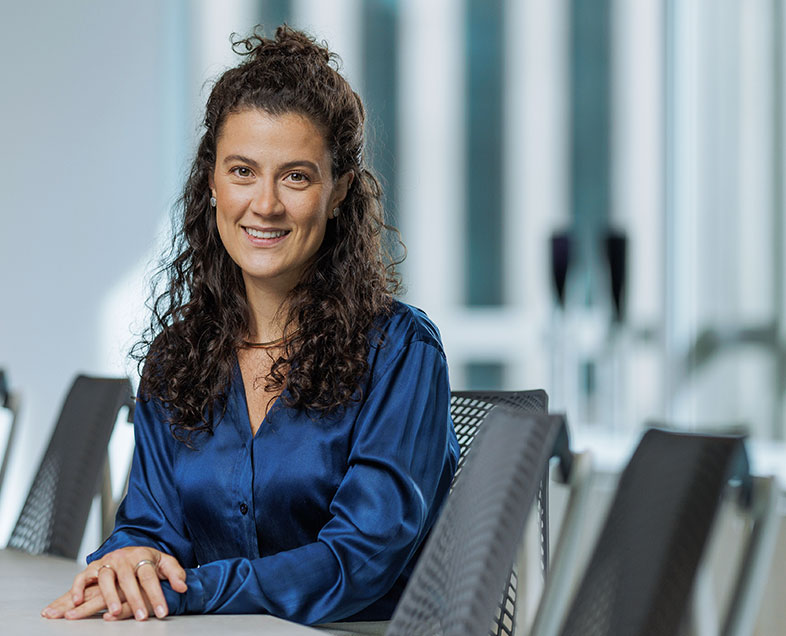 Stephanie Sartori Gennari  Mulher de cabelo preto comprido e cacheado. Veste uma blusa azul escura de mangas longas bufantes. Ela está sentada em uma cadeira, com os braços apoiados em uma mesa a sua frente e as mãos entrelaçadas, sorrindo.