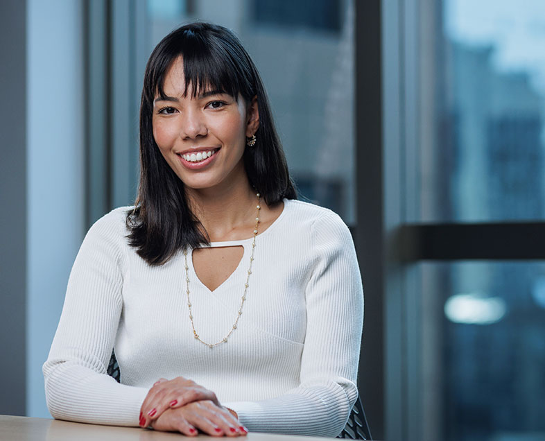 Aline Akemi Soares Kavashita  A woman with straight, shoulder-length black hair. She is wearing a long-sleeved white blouse, a long, gold, beaded necklace and her nails are painted red. She is sitting behind a desk, her hands intertwined on the table, smiling and looking straight ahead