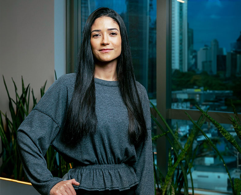 Agnes Domingues Dos Santos  Mulher de cabelos compridos lisos e pretos, está olhando para frente e com a mão direita apoiada na cintura do lado direito do corpo. Ela veste uma blusa de manga comprida com elástico na região da cintura e uma calça preta.