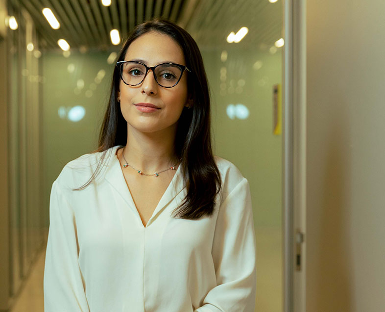 Ana Flávia Ferreira Mársico  Mulher de cabelo comprido preto. Veste uma blusa bege de mangas compridas e usa óculos. Está em pé, olhando para frente.