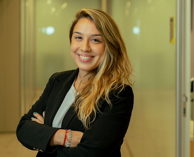 Ana Carolina Carpegiani Peyres Neves  A blonde woman with long hair. She wears a white shirt, a long-sleeved black suit and a beaded bracelet with a red ribbon on her right wrist. She is standing facing left, with her arms crossed, smiling and looking straight ahead.