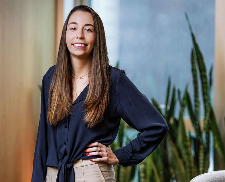 Ana Carolina Nista Gonçalves  Mulher de cabelo castanho comprido. Veste uma camisa azul escura de mangas compridas com um nó na altura da cintura, na parte da frente e uma calça bege. Ela está em pé, com a mão esquerda apoiada na cintura do lado esquerdo, está sorrindo e olhando para frente. 