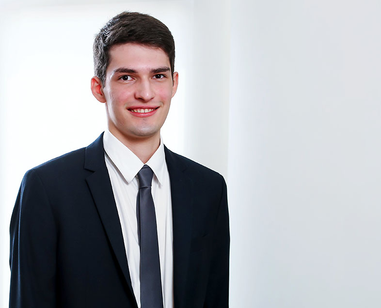 Arthur Lage  Homem de cabelo curto preto. Veste uma camisa branca, terno preto e gravata cinza escura. Ele está em pé olhando para frente e sorrindo.