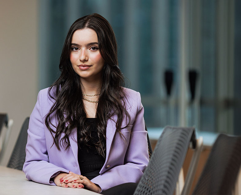 Beatriz Germano Bezerra  Mulher de cabelo preto e ondulado. Veste uma blusa preta e um blazer roxo de mangas longas. Ela esta sentada em uma cadeira, com as mãos entrelaçadas e o braço direito apoiado sobre a mesa, esta olhando para frente e sorrindo.