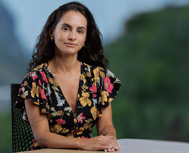 Bianca Santos Correa  Mulher de cabelos compridos castanhos e ondulados. Veste um vestido de mangas curtas com estampas de flores coloridas. Ela está sentada em uma cadeira, com as mãos entrelaçadas e apoiadas na mesa a sua frente, está sorrindo e olhando para frente.