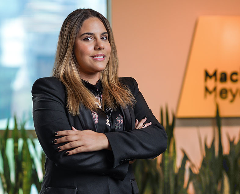 Caroline De Sena Rocha  Mulher de cabelo loiro na altura dos ombros. Veste uma blusa preta com estampas de flores cor de rosa e um blazer preto. Ela está em pé virada para a direita, de braços cruzados, sorrindo e olhando para frente.