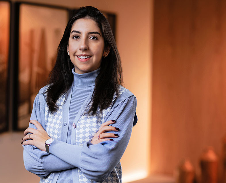 Clarissa Torrente  Mulher de cabelo preto comprido. Veste uma blusa azul de gola alta e mangas longas e um colete azul claro com estampa de ondas brancas na vertical, usa um relógio no pulso esquerdo. Ela está em pé, sorrindo e olhando para frente.