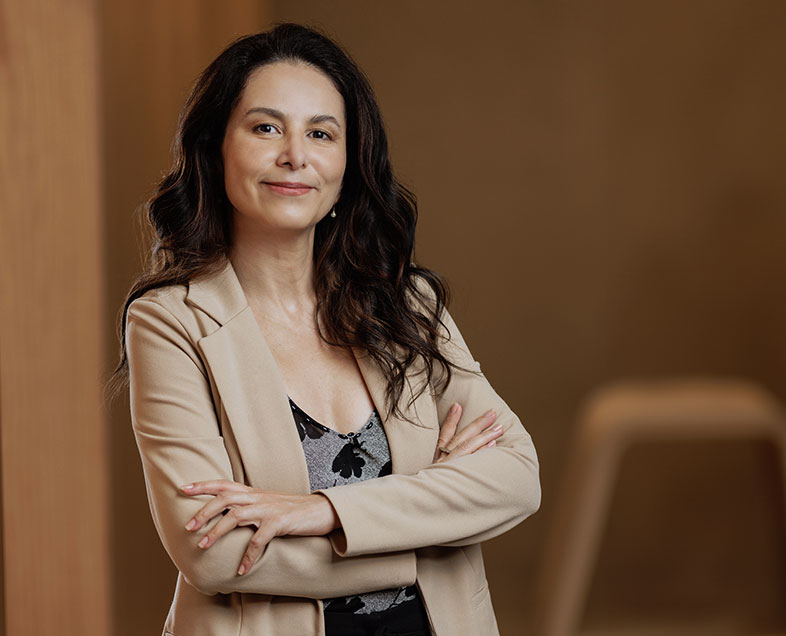 Clara Meirice Ribeiro Mendes  Mulher de cabelo castanho comprido e cacheado. Veste uma blusa cinza com estampa de flores pretas e um blazer rosa claro. Ela está em pé, de braços cruzados, olhando para frente.                                           