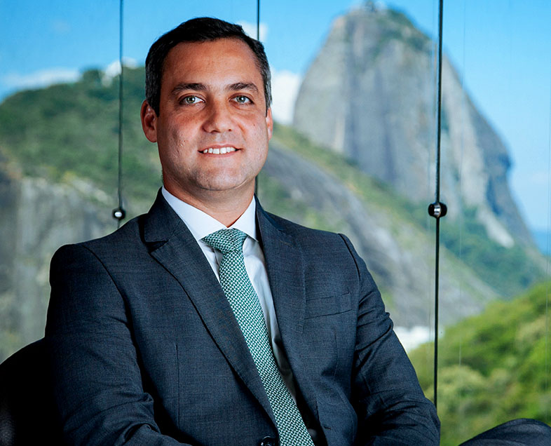 Decio Pio Borges De Castro  Homem de cabelo curto preto Veste uma camisa branca, terno cinza e gravata verde com bolinhas brancas. Ele está sentado em uma cadeira, com as pernas cruzadas, sorrindo e olhando para frente.