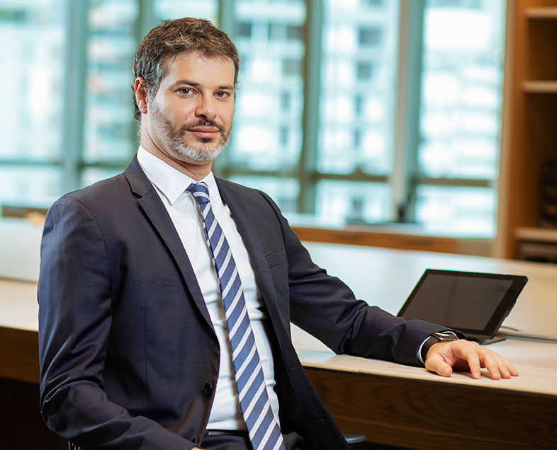 Daniel Szyfman  Homem de cabelo castanho curto, meio grisalho, com barba e bigode curtos e grisalhos. Veste uma camisa branca, terno cinza, gravata azul com listras brancas e usa relógio no pulso esquerdo. Ele está sentado em uma cadeira, virado para a direita, com o braço esquerdo apoiado em uma mesa ao seu lado, olhando para frente.