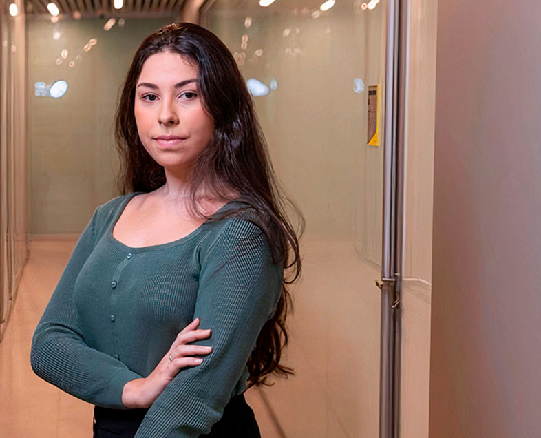 Débora Batista Matos da Silva  A woman with long black hair. She is wearing a green short-sleeved blouse. She is standing, facing to the left, with her right arm resting on her left arm, which is stretched out beside her body. 