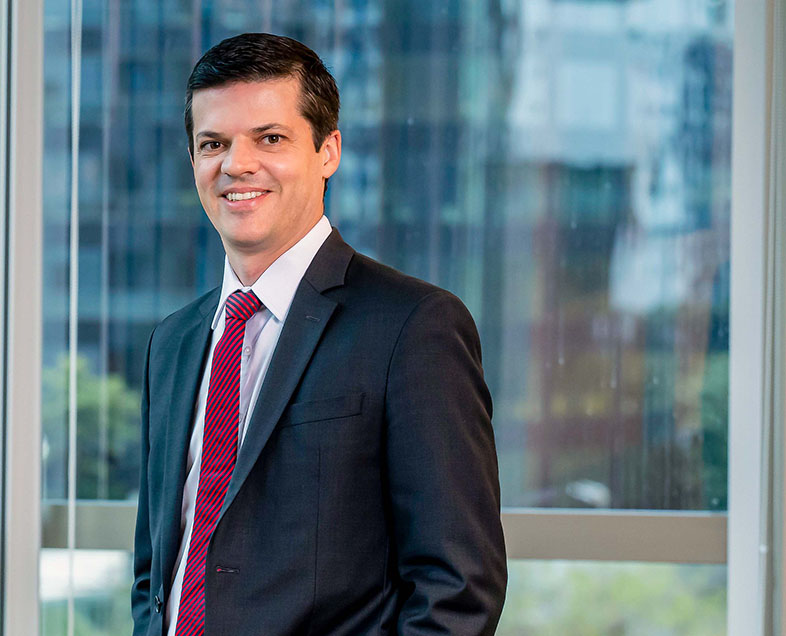 Eduardo Ferreira  A man with black hair. He's wearing a white shirt, a gray suit and a red tie with diagonal black stripes. He is standing facing left, smiling and looking straight ahead.