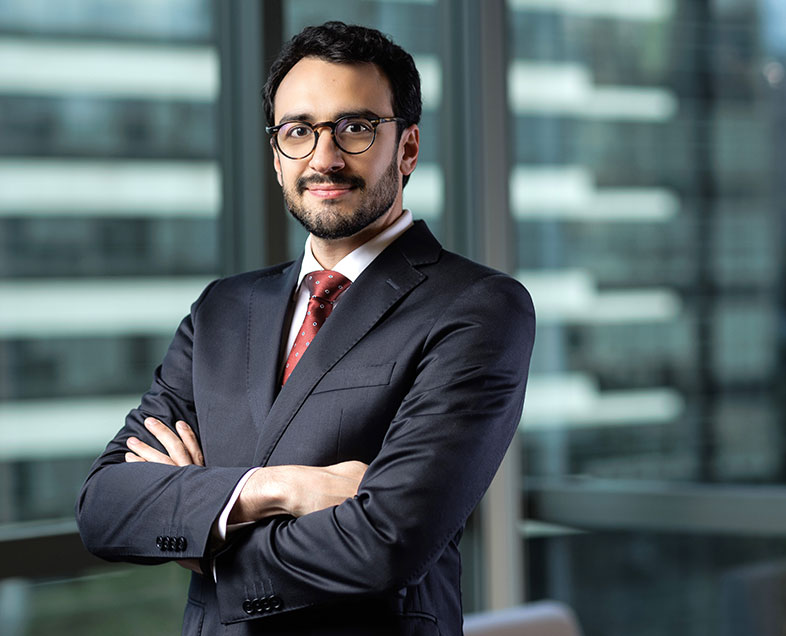 Eduardo Boldrin Camara  Homem de cabelo preto curto, com barba e bigode curtos da mesma cor. Veste uma camisa branca, terno cinza escuro, gravata vermelha e usa óculos. Ele está em pé, com os braços cruzados, olhando para frente e sorrindo. 