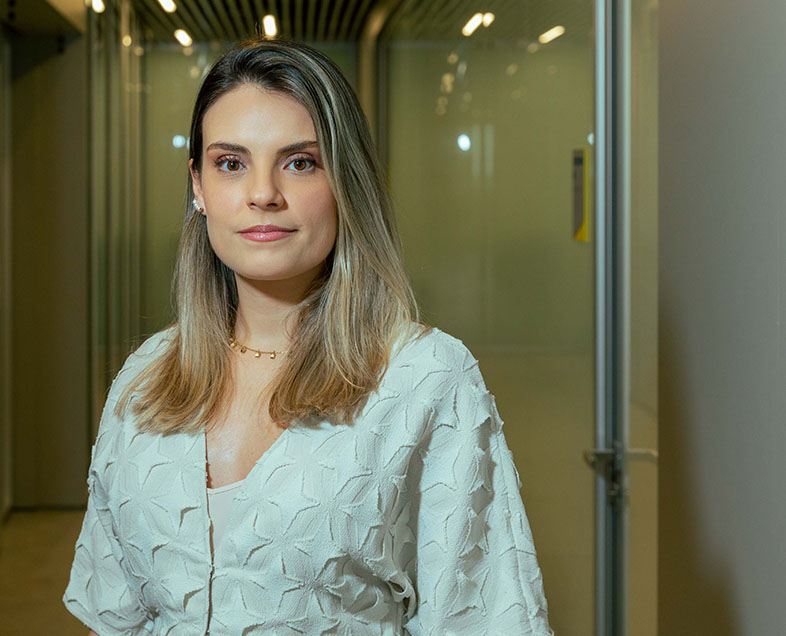 Fernanda Cury Messias  A woman with shoulder-length blonde hair. She wears a light green long-sleeved blouse with embroidered flowers in bas-relief. She is standing, looking straight ahead. 