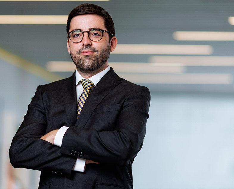 Fernando Xavier  Homem de cabelo preto curto, com barba e bigode curtos na mesma cor. Veste uma camisa branca, terno preto, gravata quadriculada preta e amarela e usa óculos. Ele está em pé, de braços cruzados e olhando para frente. 