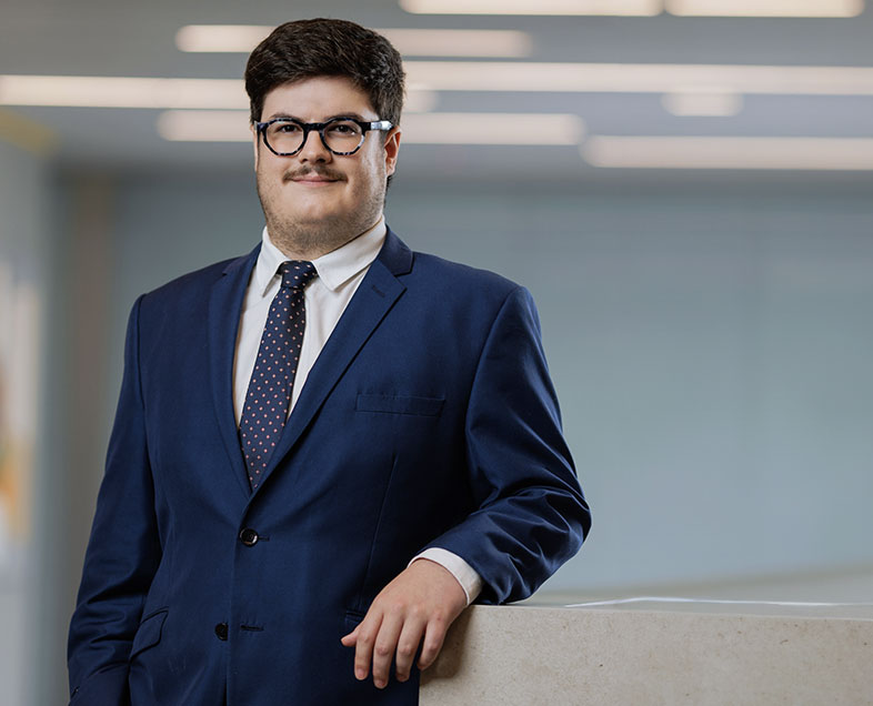 Flavio Pelone  Homem de cabelo castanho curto, com bigode curto na mesma cor. Veste uma camisa branca, terno azul escuro, gravata azul com bolinhas amarelas e usa óculos. Ele está em pé, com o braço esquerdo apoiado em uma bancada ao seu lado, está olhando para frente.