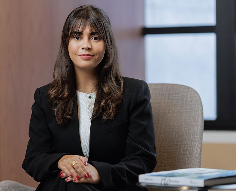 Fernanda Marques Ribeiro  Mulher de cabelo castanho comprido e ondulado. Veste uma blusa branca e um blazer preto. Ela está sentada em uma cadeira, com as pernas cruzadas e as mãos apoiadas sobre o joelho, está olhando para frente.