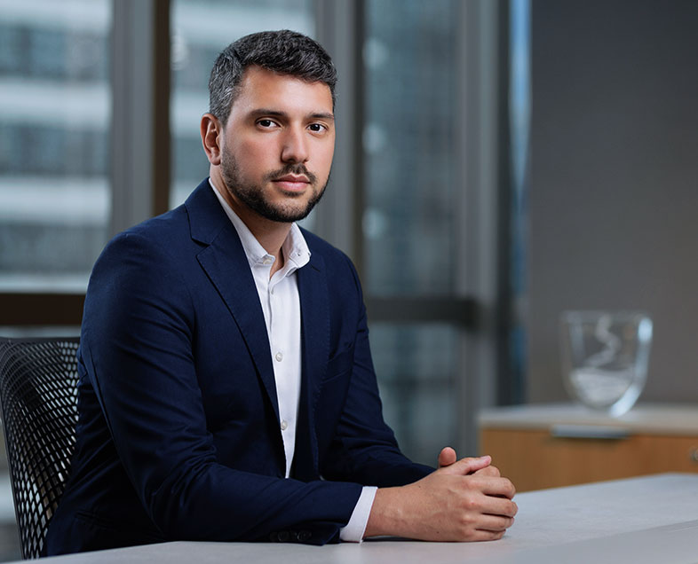 Guilherme Molledo Secco dos Santos  Homem de cabelo curto preto, com barba e bigode curtos da mesma cor. Veste uma camisa branca e blazer azul. Ele está sentado em uma cadeira, virado para a direita, com as mãos entrelaçadas e apoiadas em uma mesa a sua frente.