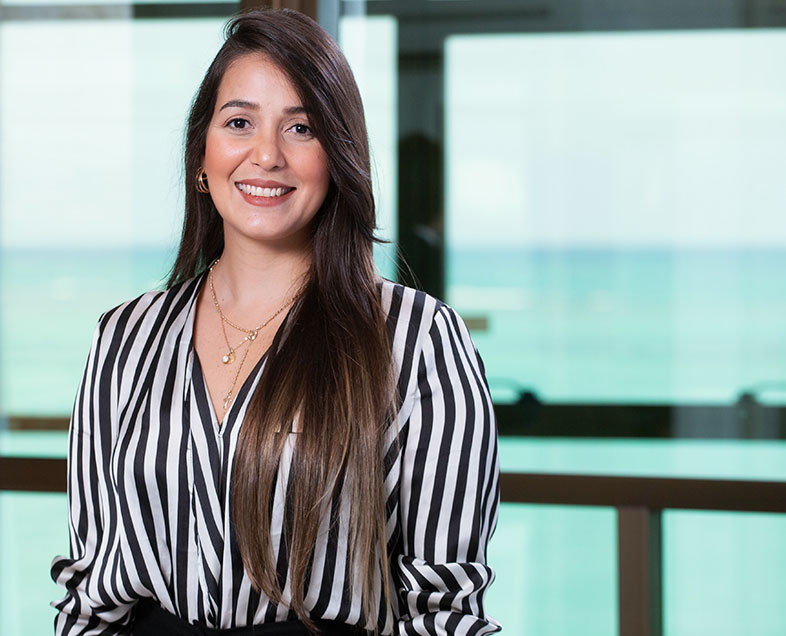 Gabriella Maria Cavalcanti do Nascimento  Mulher de cabelo castanho comprido. Veste uma blusa de mangas longas branca com estampa de listras de pretas na vertical. Ela está em pé, olhando para frente e sorrindo.