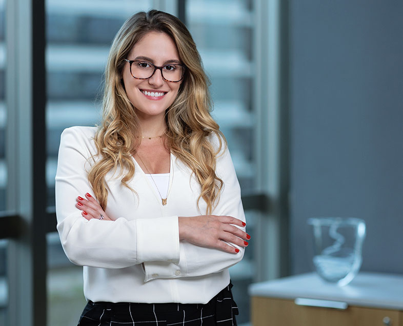 Giovanna Pasquini Malfatti  Mulher de cabelo loiro comprido e ondulado. Veste uma blusa branca de mangas compridas e usa óculos. Ela está em pé, de braços cruzados e sorrindo.