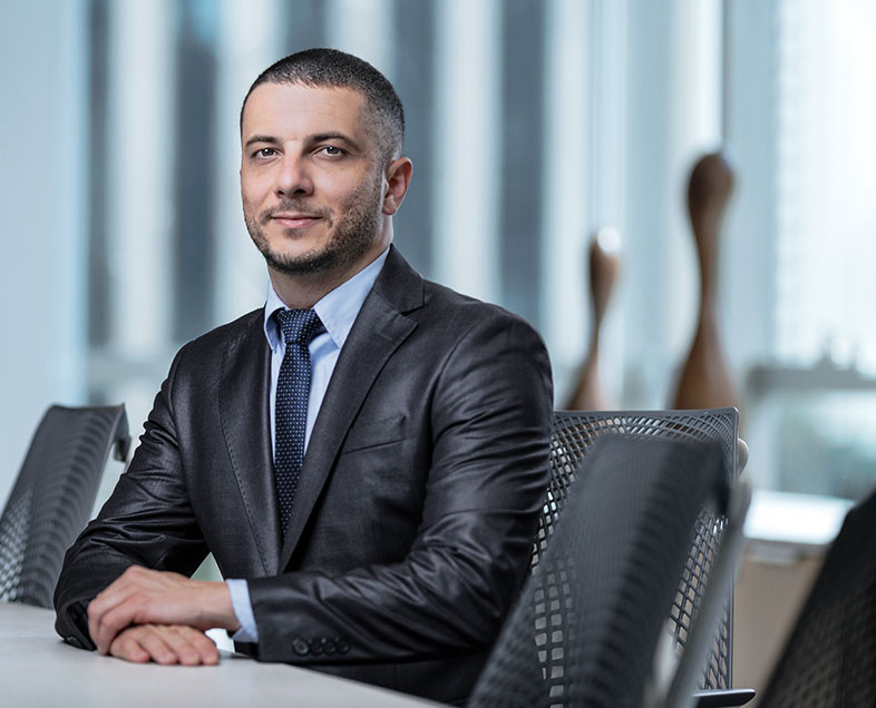 Gabriel de Castro Dias  Homem de cabelo curto preto, com barba e bigode curtos. Veste uma camisa azul, terno preto e gravata preta. Ele está sentado em uma cadeira, com as mãos apoiadas em uma mesa a esquerda, está olhando para frente.