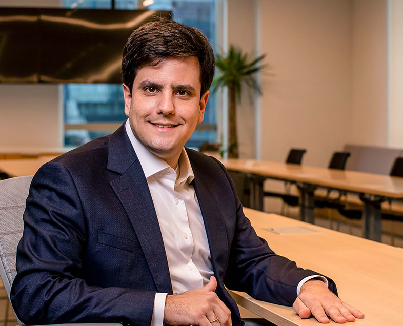 Gustavo Secaf Rebello  Homem de cabelo castanho curto. Veste uma camisa branca e um blazer azul. Ele está sentado em uma cadeira, com o braço direito apoiado no encosto da própria cadeira e o braço esquerdo apoiado em uma mesa ao seu lado, está sorrindo. 