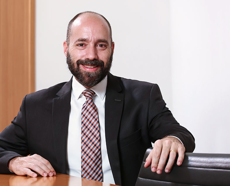 Gustavo Rugani  Homem de cabelo preto, quase calvo, com barba e bigode medianos da mesma cor. Veste uma camisa branca, terno preto e gravata com estampa quadriculada preta, laranja e azul. Ele está sentado em uma cadeira, com o braço esquerdo apoiado no encosto de uma cadeira ao seu lado e o braço direito apoiado na mesa a sua frente, está sorrindo.