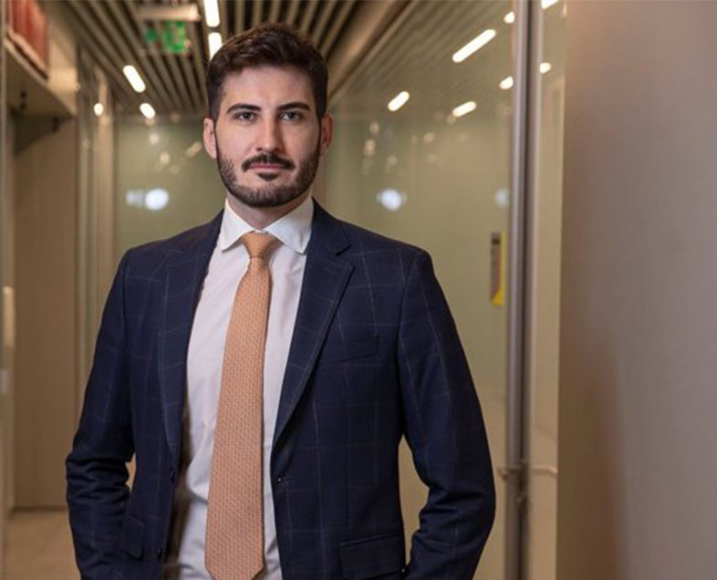 Guilherme Spinacé  Homem de cabelo curto preto, com barba e bigode curtos da mesma cor. Veste uma camisa branca, terno azul escuro e gravata laranja claro. Ele está em pé, com as mãos nos bolsos da calça e olhando para frente.