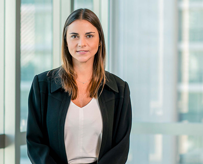 Diana Henne  Mulher de cabelo loiro na altura dos ombros, vestindo uma blusa branca e um blazer preto. Ela está em pé, olhando para frente e sorrindo.