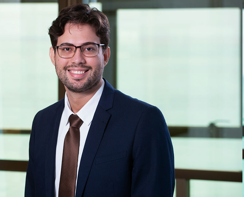 Heitor Lima Gonçalves de Oliveira  Homem de cabelo castanho curto e cacheado, com barba e bigode curtos da mesma cor. Veste uma camisa branca, terno azul escuro, gravata marrom e usa óculos. Ele está em pé, sorrindo.