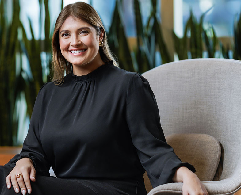 Helena Prescinotti Cursino de Andrade  Mulher de cabelo castanho comprido. Veste uma blusa preta de mangas longas. Ela está sentada em uma cadeira, virada para a esquerda, com as pernas cruzadas e o braço direito apoiado sobre elas, está sorrindo e olhando para frente.