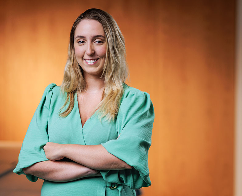 Isabelle Valente  Mulher de cabelo loiro na altura dos ombros. Veste uma blusa verde clara de mangas curtas e largas. Ela está em pé, de braços cruzados e sorrindo.