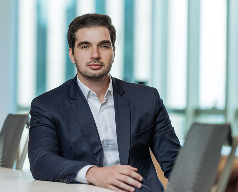 Ivan Fogaça Panza  Homem de cabelo curto preto, com barba e bigode curtos da mesma cor. Veste uma camisa branca e um terno cinza escuro. Ele está sentado em uma cadeira, com o braço esquerdo apoiado na mesa ao seu lado, olhando para frente.