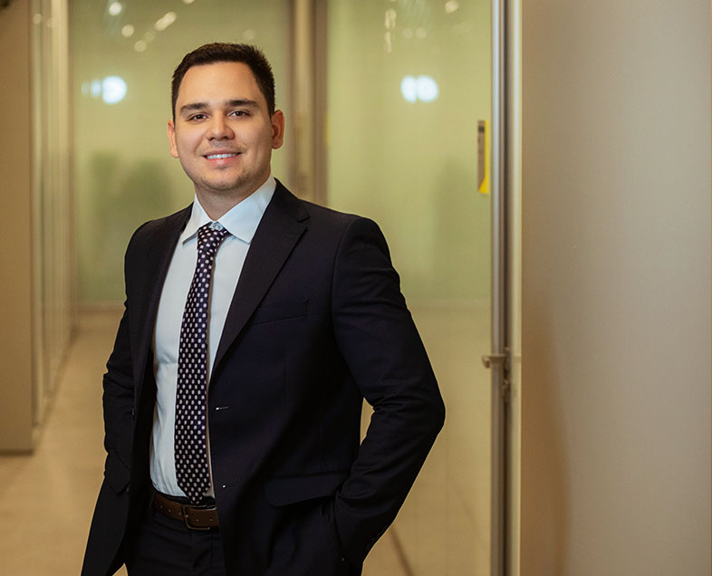 Joao Pedro Gargantini Grapella Leite  A man with short black hair. He's wearing a white shirt, black suit, black pants and a black tie with white polka dots. He is standing with his hands in his trouser pockets, smiling.