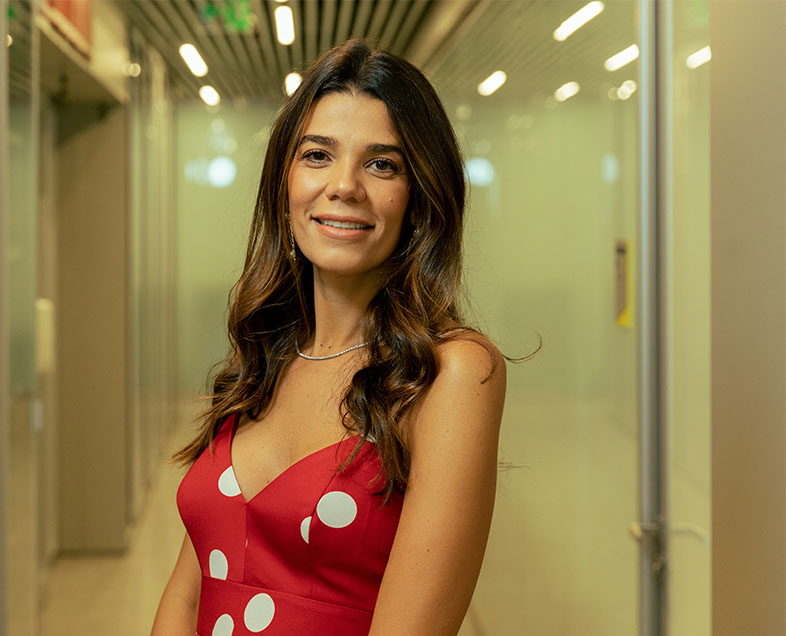 Juliana Caleiro  Mulher de cabelo castanho comprido. Veste um vestido de alças, vermelho com bolinhas brancas. Ela está em pé, virada para a esquerda, olhando para frente e sorrindo.