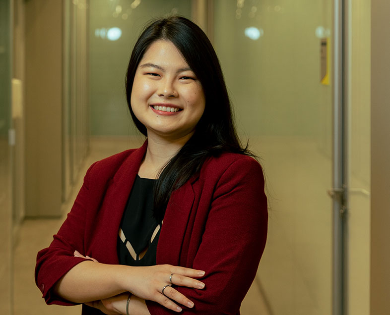 Jéssica Kaori Konishi   Mulher de cabelo preto comprido. Veste uma blusa preta e um blazer vermelho de meia manga. Ela está em pé, de braços cruzados e sorrindo.