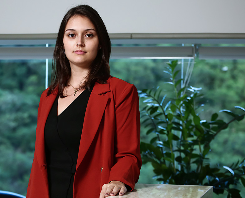 Larissa Barreto Vadnjal Moraes  Mulher de cabelo preto na altura dos ombros. Veste uma blusa preta e um casaco vermelho. Ela está em pé, com a mão esquerda apoiada em uma bancada, olhando para frente.