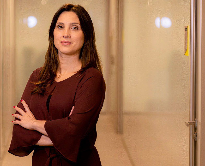 Luciana Costa Engelberg  Mulher de cabelo castanho comprido. Veste um vestido vinho com uma abertura na vertical na altura do peito e com mangas largas na altura dos cotovelos. Ela está em pé, de braços cruzados, olhando para frente.   