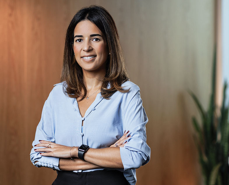 Ligia dos Santos de Andrade  Mulher de cabelo castanho curto. Veste uma camisa azul clara, calça preta e usa relógio no pulso esquerdo. Ela está em pé, de braços cruzados e sorrindo.