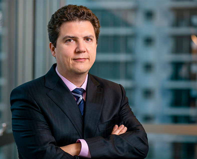 José Virgílio Lopes Enei  A man with short, curly brown hair. He wears a light pink shirt, a black suit and a dark blue tie. He is standing with his arms crossed, looking straight ahead.