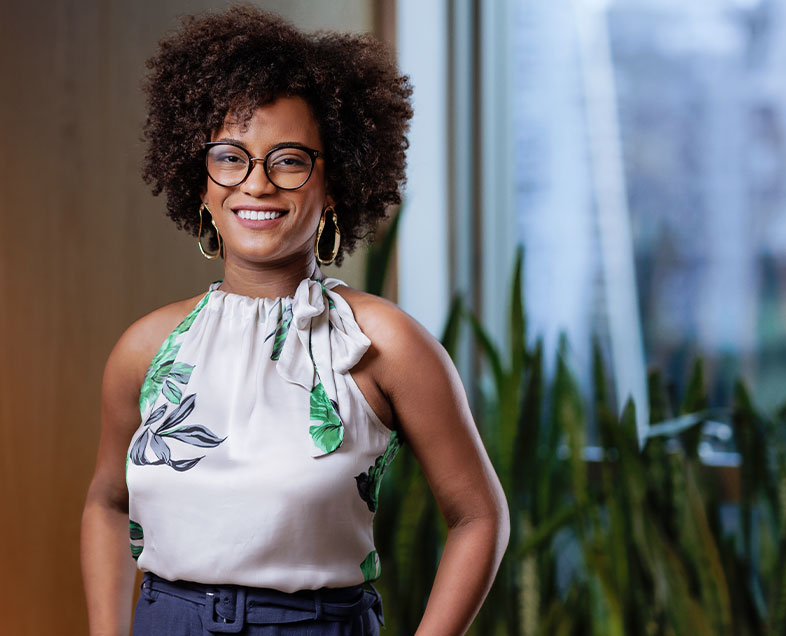 Lígia de Cássia Pereira Silva Pinto  Mulher de cabelo castanho mediano e cacheado. Veste uma blusa cinza clara, sem mangas, com estampa de folhas verdes e azul e um laço no pescoço do lado esquerdo, calça azul e usa óculos. Ela está em pé, sorrindo.