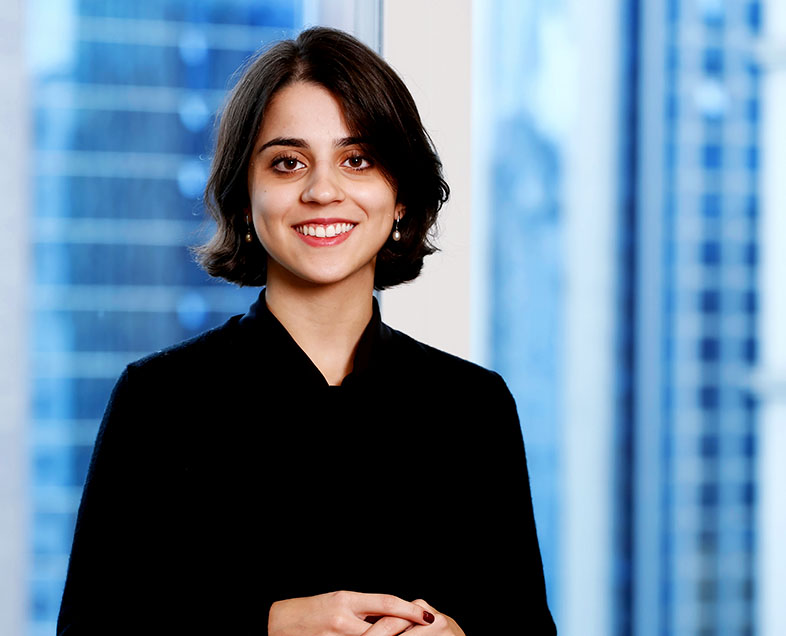 Larissa Gebrim  A woman with short brown hair. She is wearing a long-sleeved black shirt. She is standing with her hands clasped at her chest, looking straight ahead and smiling.