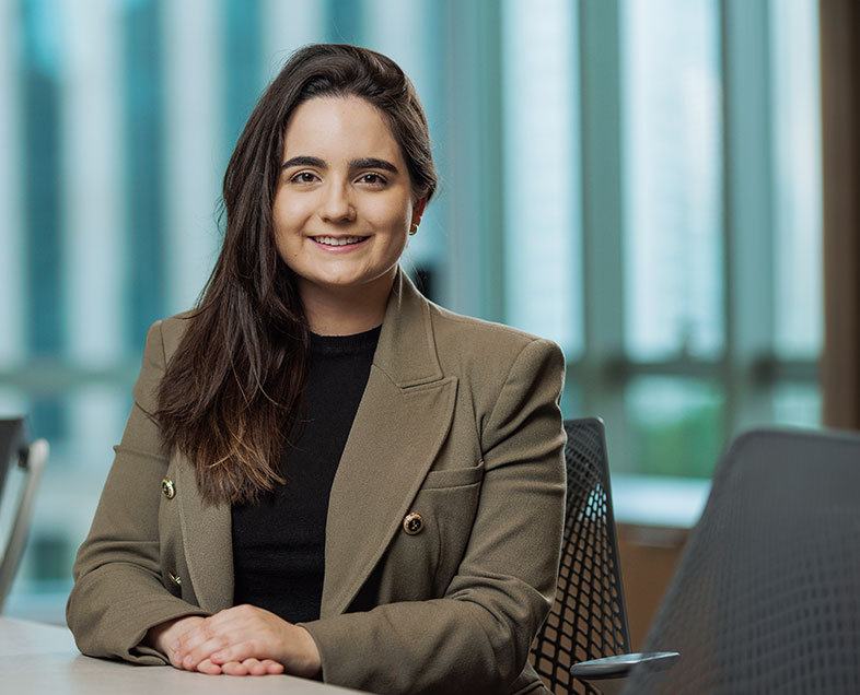 Luiza Toscano   Mulher de cabelo castanho comprido. Veste uma blusa preta com um blazer cor caqui. Ela está sentada em uma cadeira, com as mãos entrelaçadas e apoiadas em uma mesa a sua frente, sorrindo.