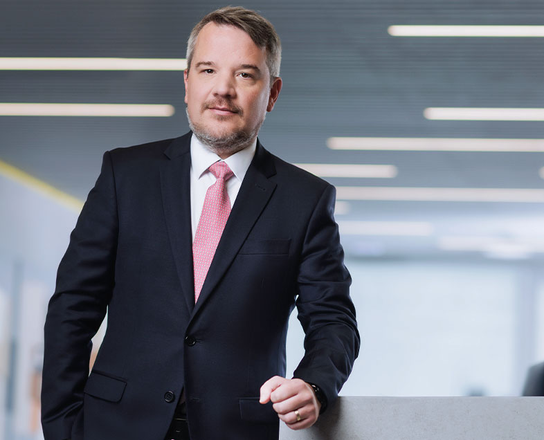 Marco Behrndt  A man with short brown hair, a short beard and mustache of the same color. He is wearing a white shirt, black suit and red tie. He is standing with his left arm resting on a bench beside him.