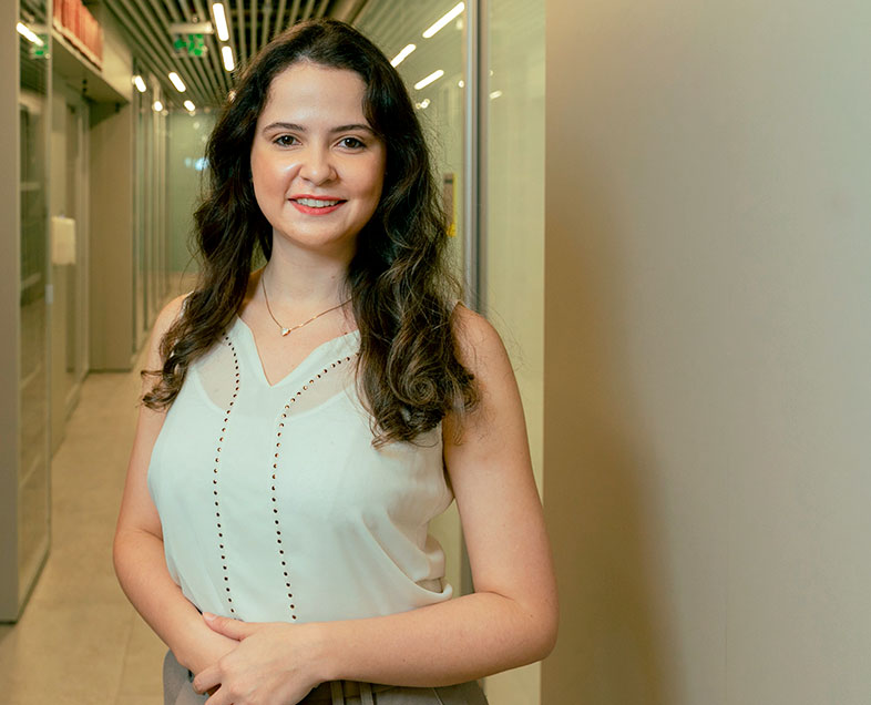 Maria Cecilia Da Conceicao Faria Dos Santos  A woman with long brown hair. She is wearing a light green sleeveless blouse. She is standing with her hands clasped at her belly, smiling.