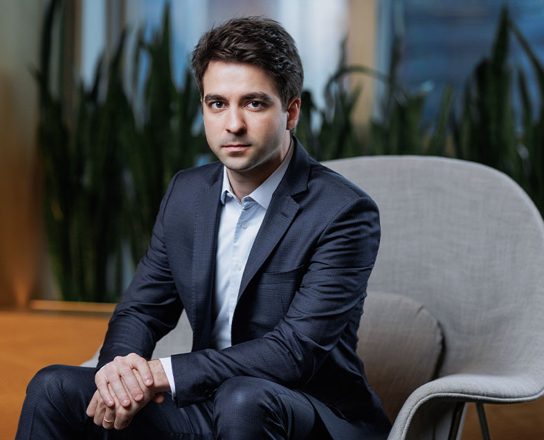 Murilo Caldeira Germiniani  A man with short brown hair. He is wearing a light blue shirt and a dark blue suit. He is sitting in an armchair with his arms resting on his legs and looking ahead.