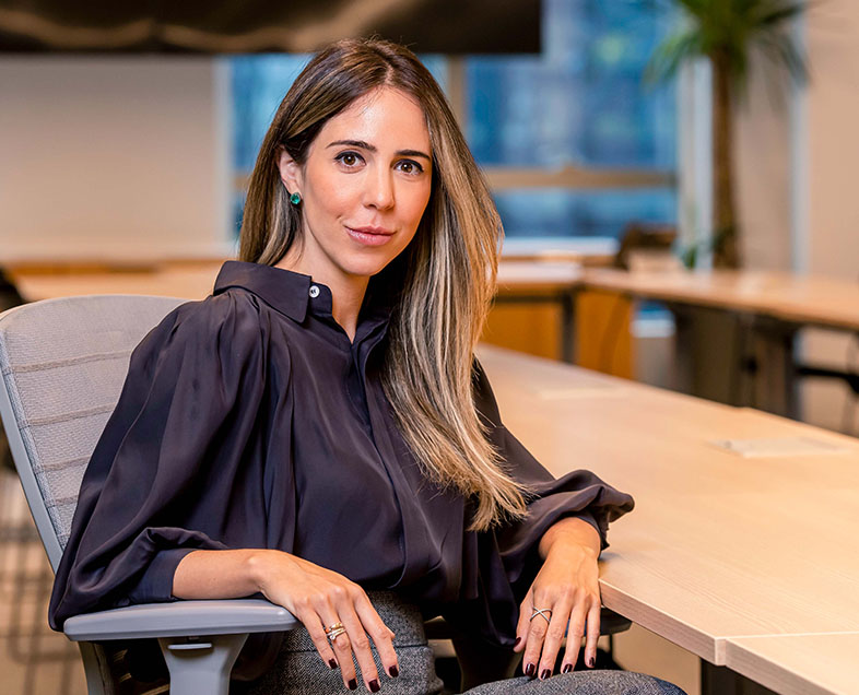 Marilia de Prince Rasi Faustino  Mulher de cabelo castanho comprido. Veste uma camisa preta de mangas compridas e uma calça cinza. Ela está sentada em uma cadeira, com o braço esquerdo apoiado na mesa ao seu lado e o braço direito apoiado no braço da cadeira, olhando para frente.