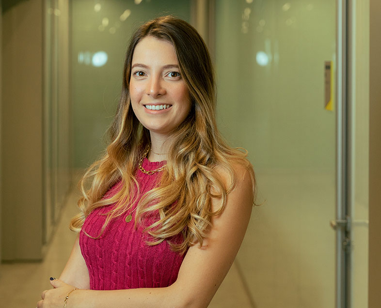 Marina Piccolotto Nori  Mulher de cabelo loiro comprido. Veste uma blusa rosa sem mangas. Ela está em pé, de braços cruzados, olhando para frente e sorrindo.