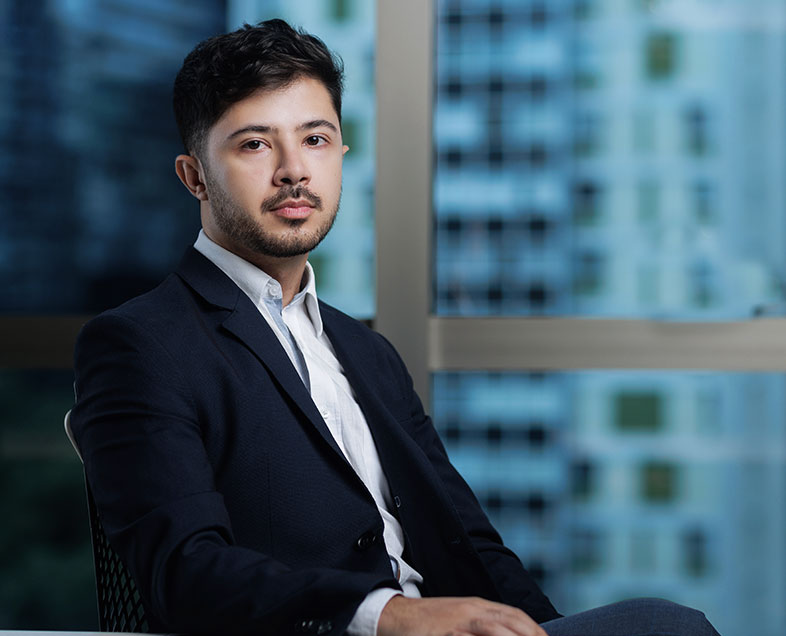 Matheus Faria de Sousa  Homem de cabelo preto curto, com barba e bigode curtos da mesma cor. Veste uma camisa branca e terno preto. Ele está sentado em uma cadeira, virado para a direita, com o braço direito apoiado em uma mesa. olhando para frente.