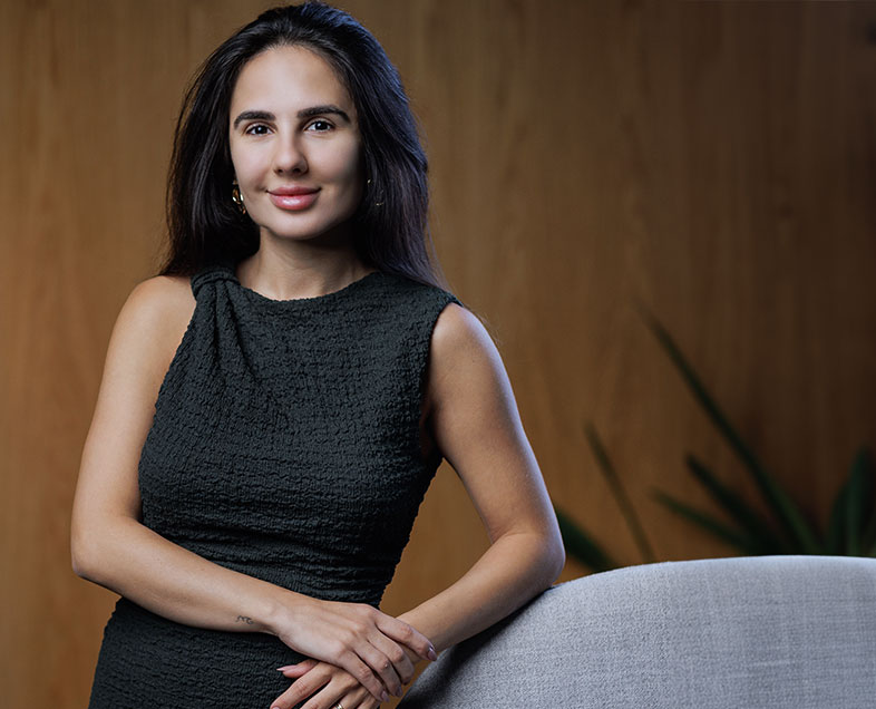 Marianna Torello Palladino  A woman with black hair tied up in a bun with a few loose strands. She is wearing a long black dress with long sleeves and a high collar. She is sitting in an armchair facing left, with her legs crossed and her hands resting on her knees, looking straight ahead and smiling. 